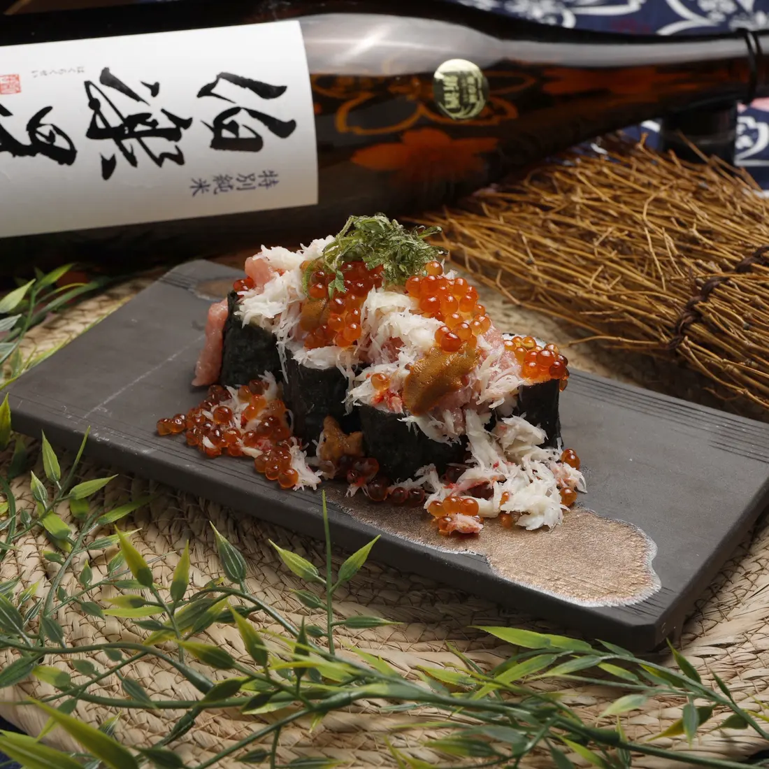 日本酒の瓶の前に置かれた、カニといくらをのせた海苔巻きの料理写真